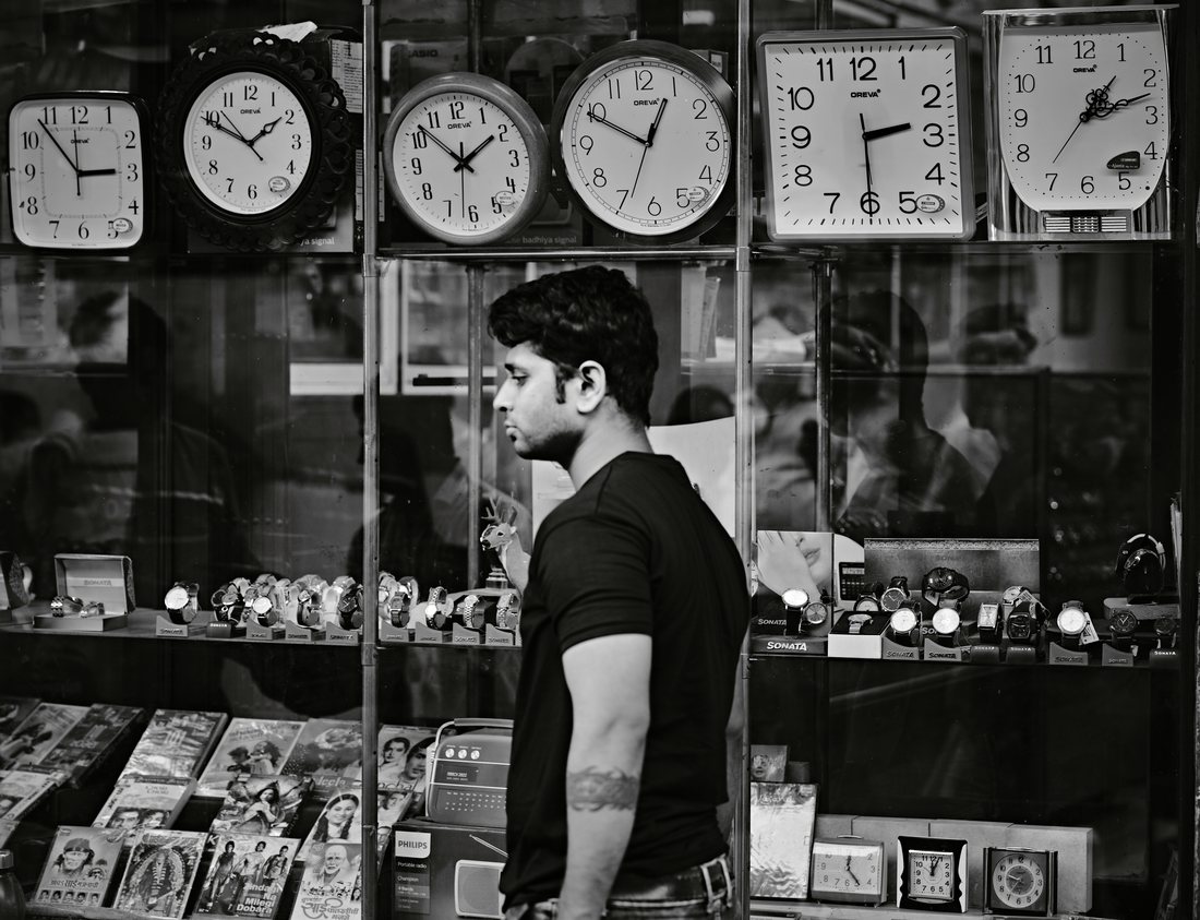 Shop owner glancing sideways beside a wall of clocks, monochrome